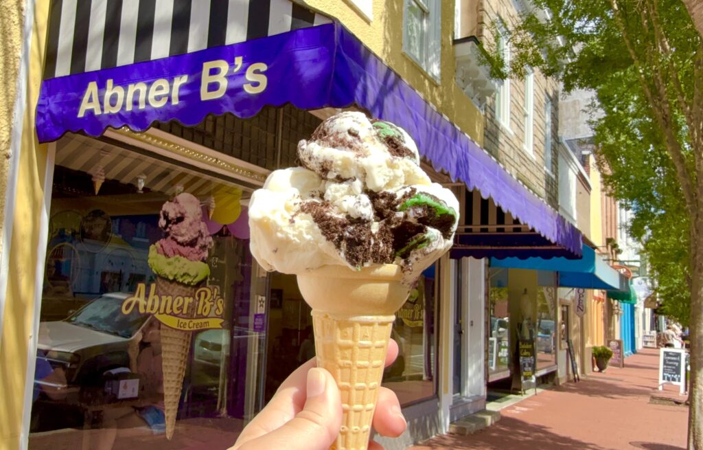 A hand holds an ice cream cone with cookies and cream flavor in front of the purple awning of Abner B&rsquo;s Ice Cream shop
