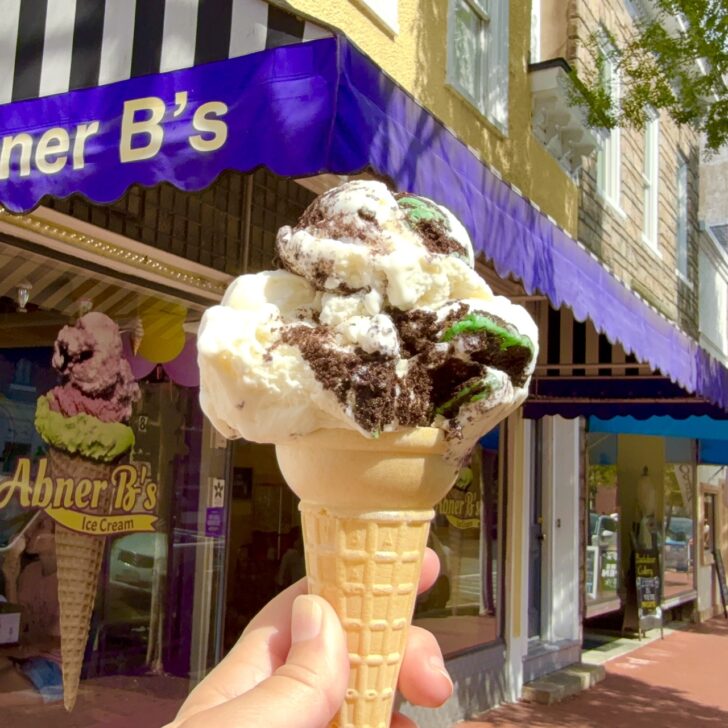 A hand holds an ice cream cone with cookies and cream flavor in front of the purple awning of Abner B’s Ice Cream shop