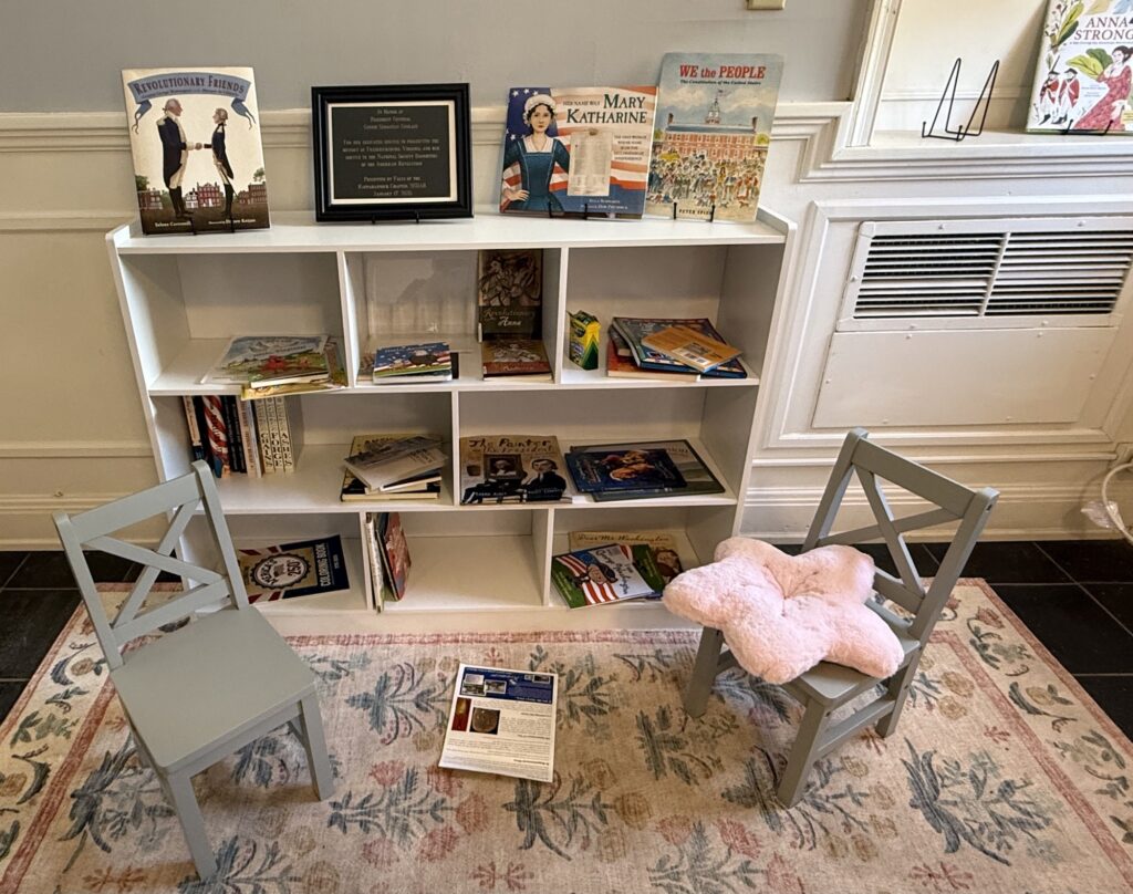 A small bookshelf filled with children's books sits against a wall, flanked by two gray chairs on a patterned rug&mdash;an inviting spot for storytime and one of the cozy things to do in Fredericksburg with kids. A pink star-shaped pillow and paper rest on the floor.