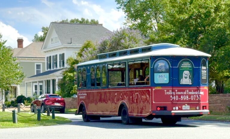 A red trolley bus labeled "Trolley Tours of Fredericksburg" drives past houses on a sunny day, making it a fun option among things to do in Fredericksburg with kids, with a red car parked nearby.
