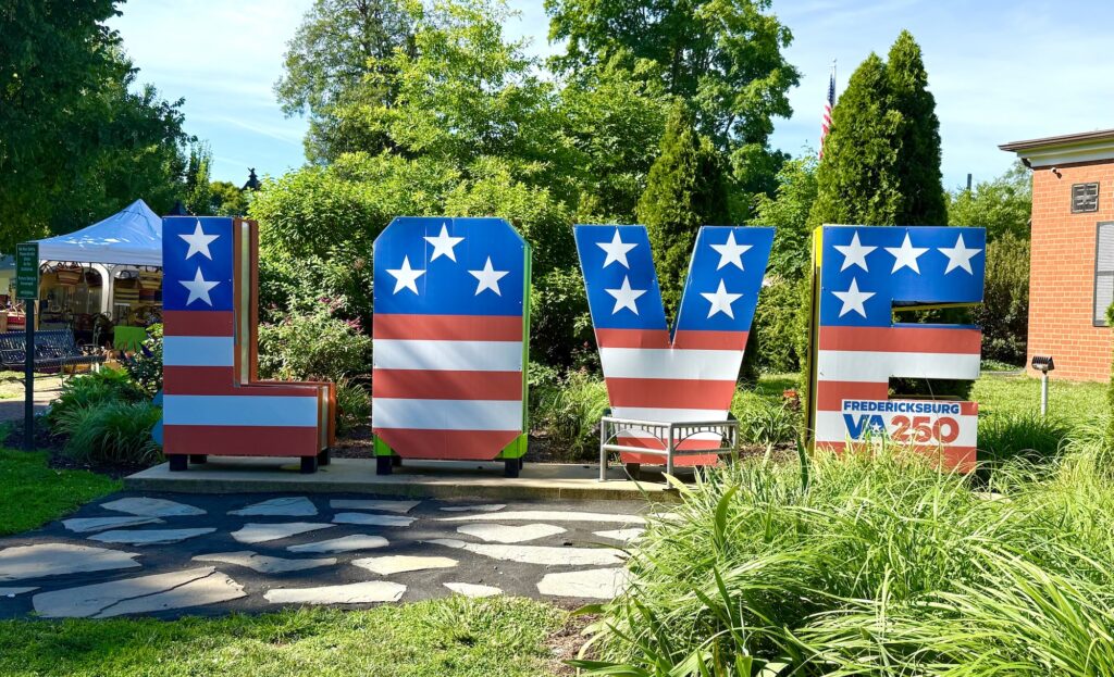 Large letters spelling "LOVE" are decorated with stars and stripes, resembling the American flag, in an outdoor setting with greenery and a Fredericksburg VA250 sign&mdash;perfect for photos and one of the best things to do in Fredericksburg with kids.