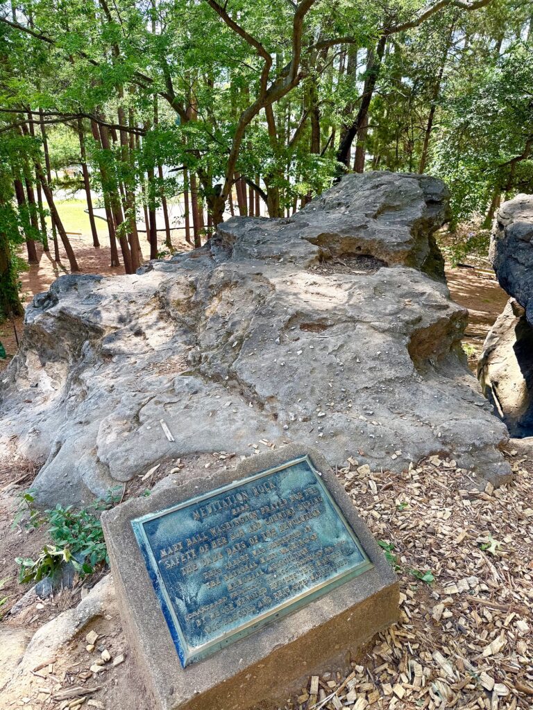 Large rock formation in a wooded park area with a metal historical plaque mounted on a concrete base&mdash;one of the unique things to do in Fredericksburg with kids who love exploring history and nature.