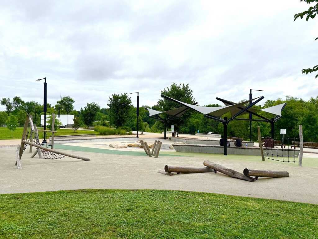 An outdoor playground featuring wooden climbing structures, rope nets, and shaded seating areas surrounded by green grass and trees&mdash;perfect for families seeking things to do in Fredericksburg with kids under a cloudy sky.