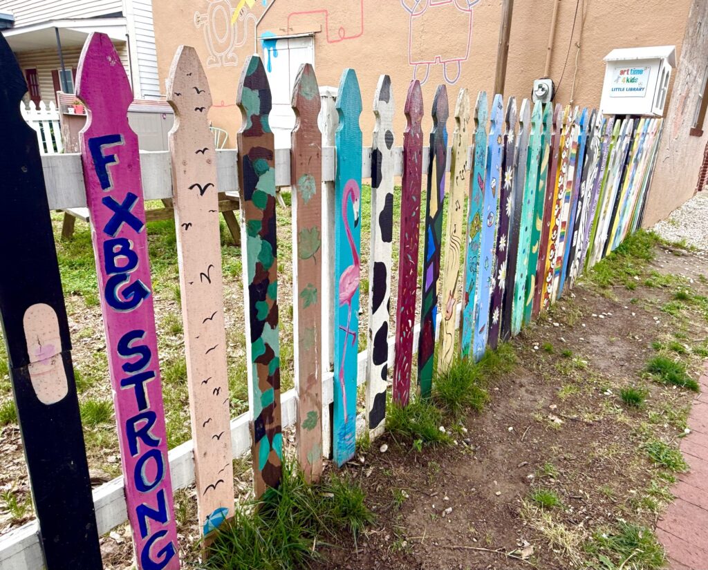 A white picket fence with each plank uniquely painted in vibrant colors, text, animals, and abstract designs outside a tan building&mdash;a fun photo spot among the best things to do in Fredericksburg with kids.