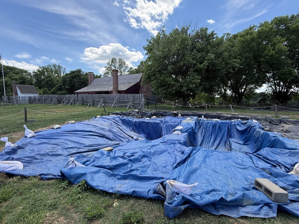 A large blue tarp covers an excavated pit, held down by sandbags and bricks, with historic buildings and trees in the background&mdash;a unique scene among the things to do in Fredericksburg with kids under a partly cloudy sky.
