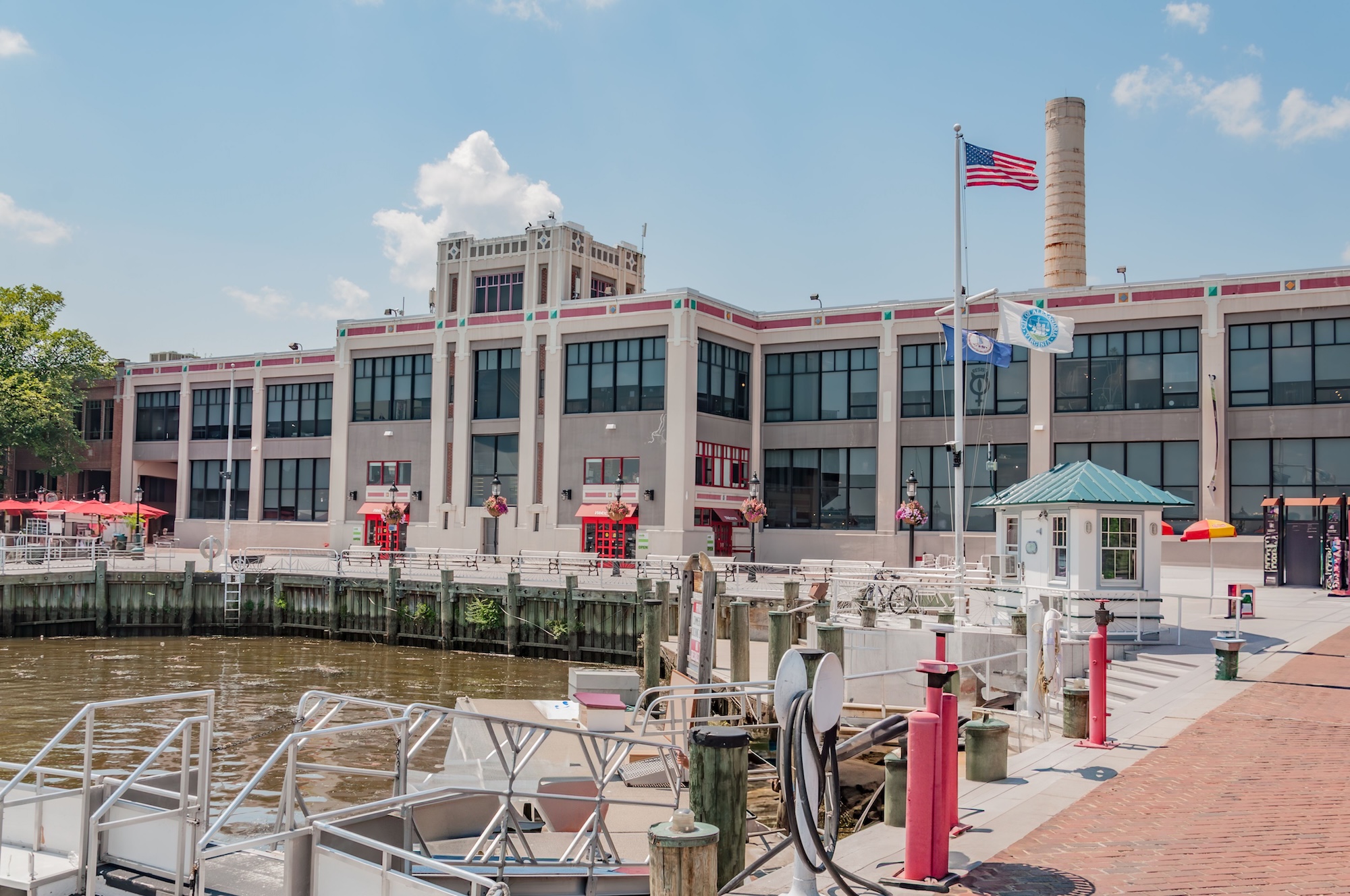 A waterfront building in Alexandria with American and state flags, docks, and boats in the foreground under a clear sky.