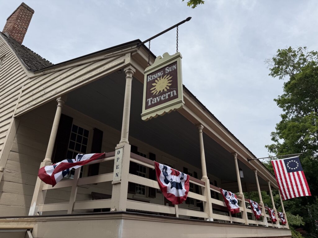 Historic building in Fredericksburg with a &ldquo;Rising Sun Tavern&rdquo; sign, decorated with red, white, and blue bunting and a Betsy Ross American flag, under a cloudy sky.