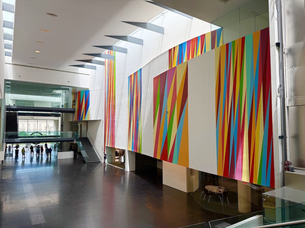 A spacious modern atrium with glass walls, colorful geometric murals, black tile floor, and a staircase welcomes visitors—making it an inviting spot for those seeking unique Virginia weekend getaways—as people walk near the entrance.