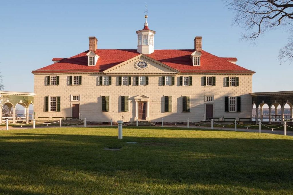 A large historic mansion with a red roof, white siding, and green shutters stands on a lawn under a clear blue sky in Alexandria.
