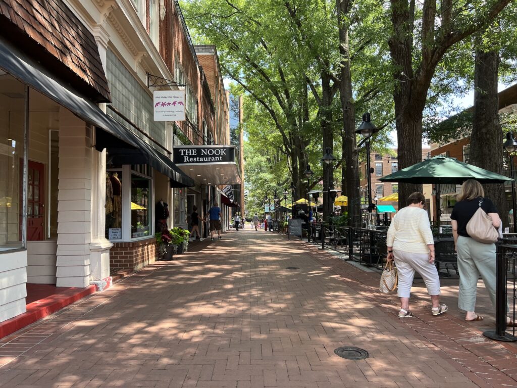 Brick-paved pedestrian street lined with shops, restaurants in Charlottesville, outdoor seating, and people walking under leafy trees on a sunny day.