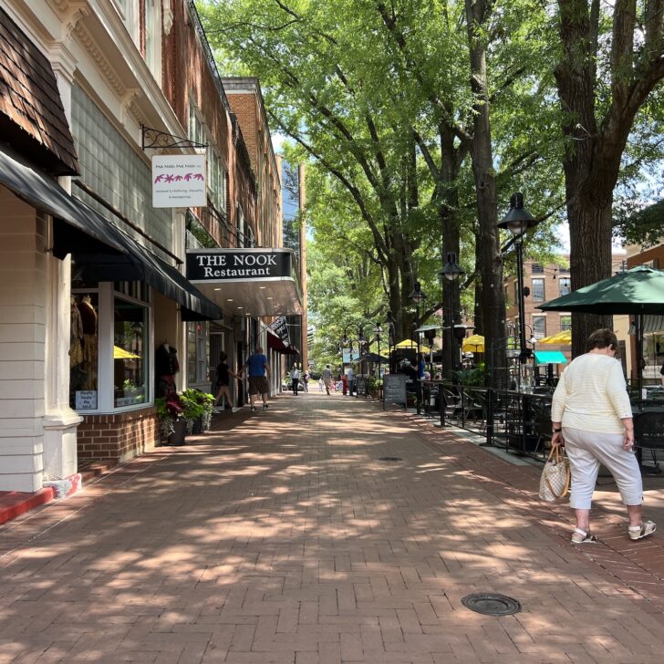 Brick-paved pedestrian street lined with shops, restaurants in Charlottesville, outdoor seating, and people walking under leafy trees on a sunny day.