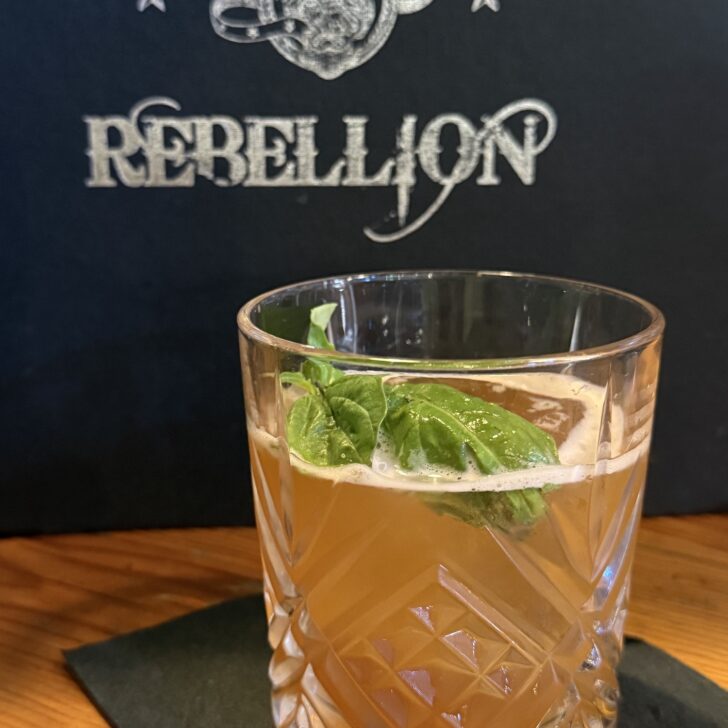 A glass of amber cocktail garnished with basil sits on a black napkin, with a “REBELLION” sign and logo visible in the background.