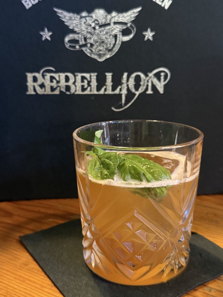 A glass of amber cocktail garnished with basil sits on a black napkin, with a &ldquo;REBELLION&rdquo; sign and logo visible in the background.