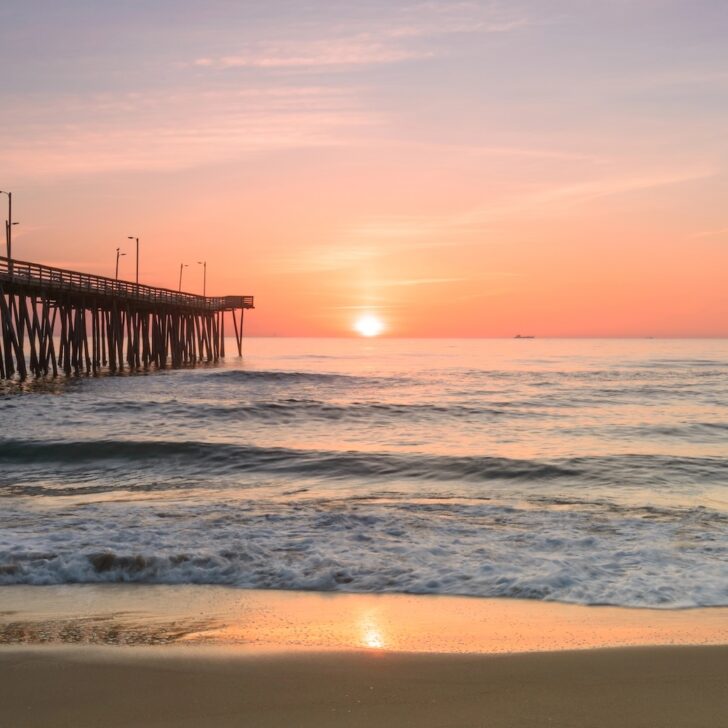A wooden pier extends into the ocean at sunrise, with gentle waves and a pastel-colored sky reflected on the water and wet sand.