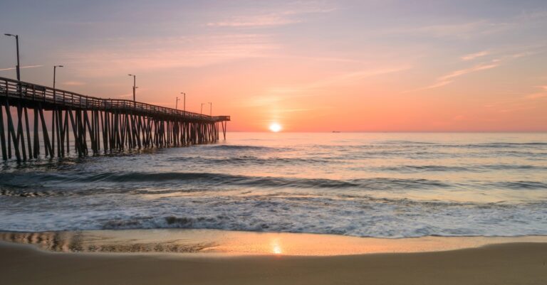 A wooden pier extends into the ocean at sunrise, with gentle waves and a pastel-colored sky reflected on the water and wet sand.