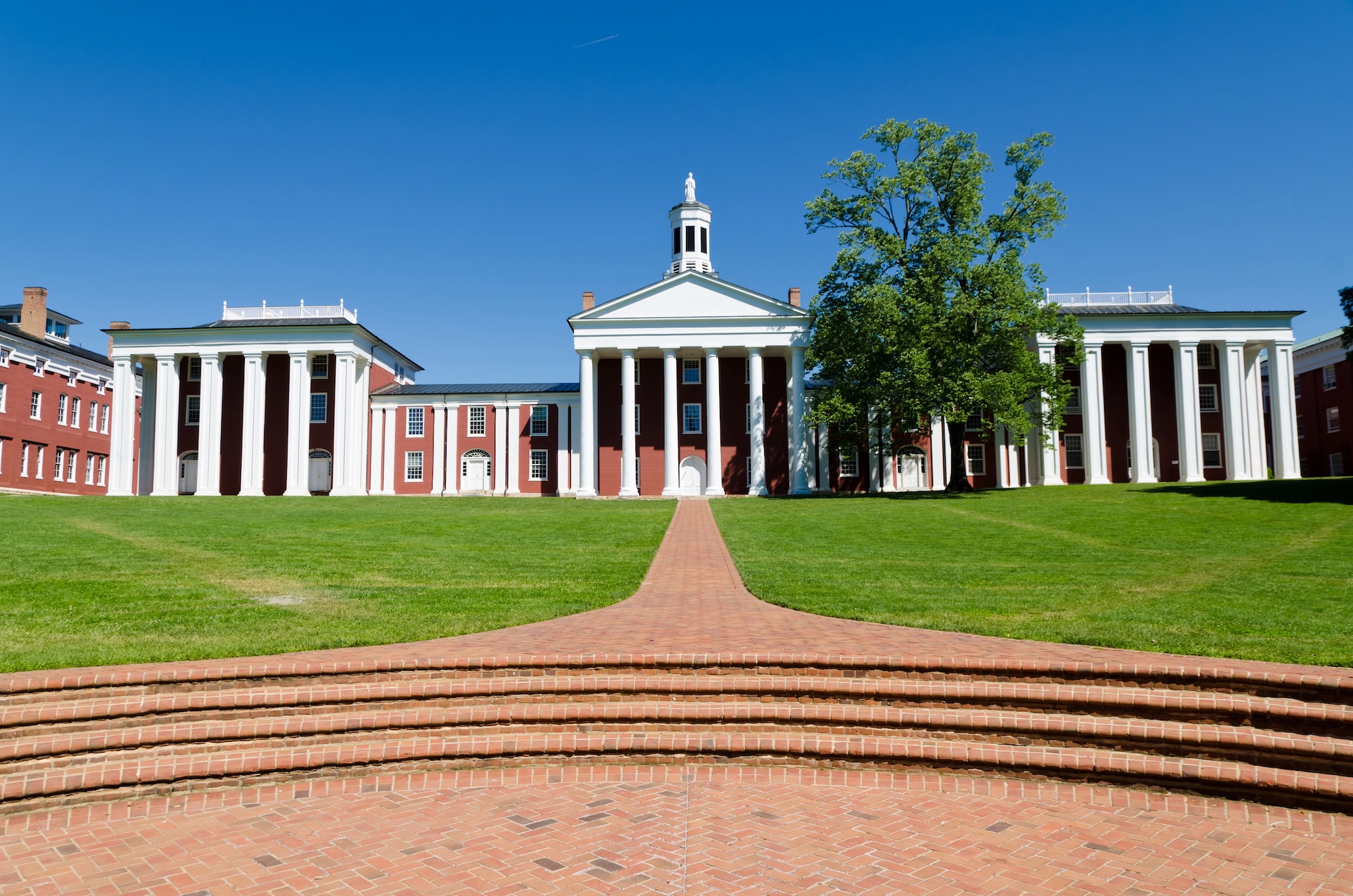 A red brick building with white columns and a central cupola sits on a grass lawn under a clear blue sky, with brick steps leading to the entrance&mdash;a classic scene often found in small towns in Virginia.