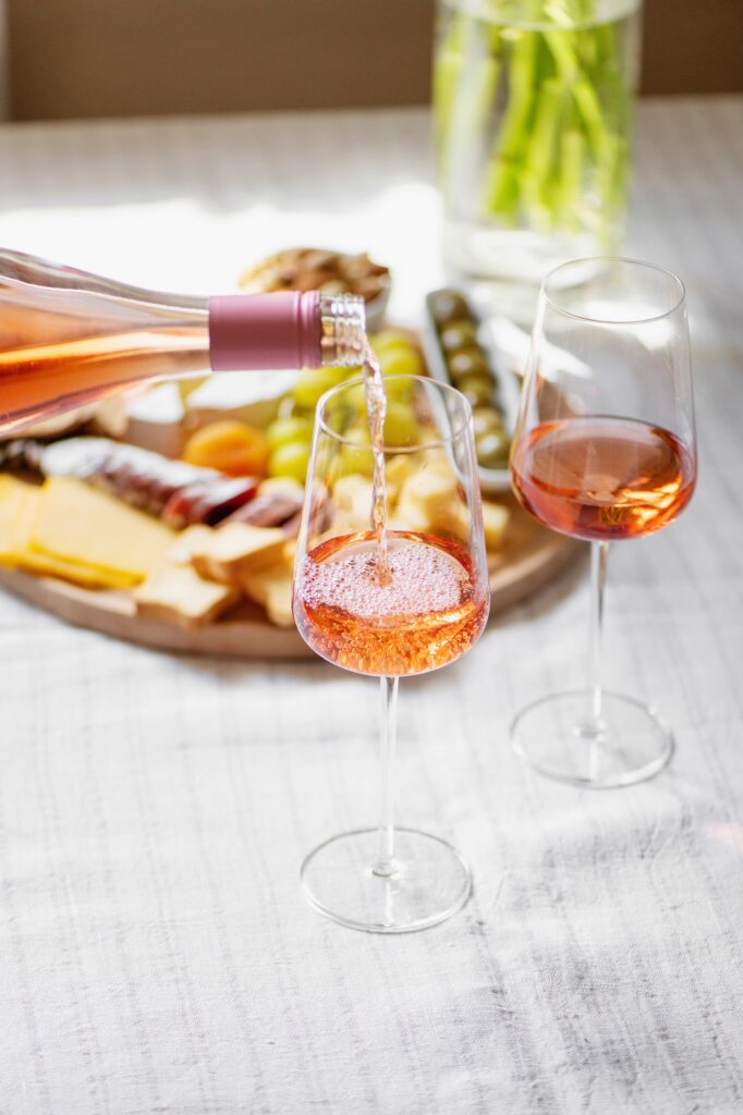 Ros&eacute; wine being poured into a glass beside another filled glass, with a cheese and charcuterie platter in the background on a light tablecloth.