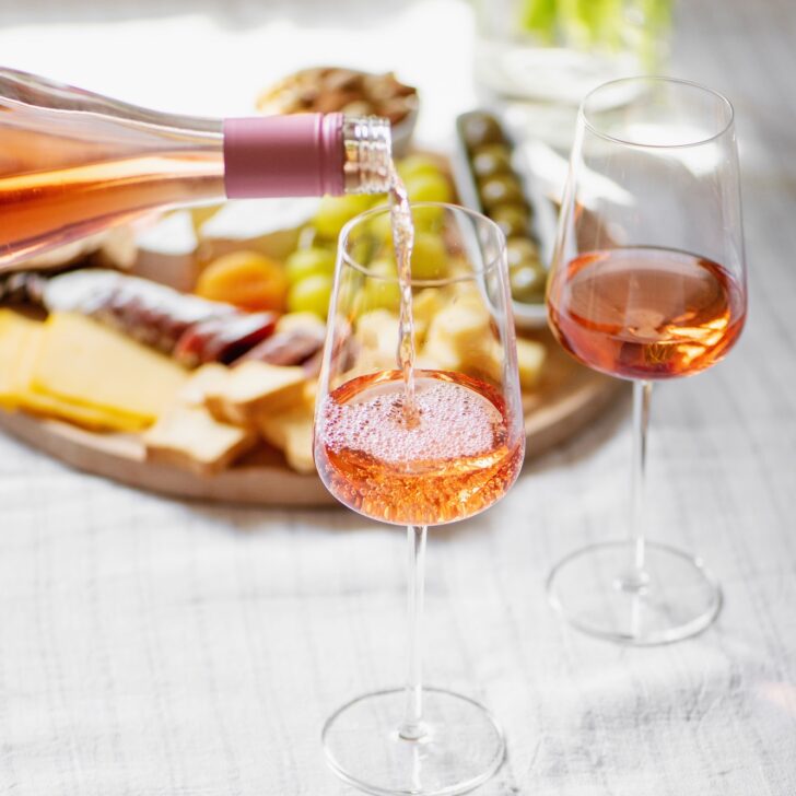 Rosé wine being poured into a glass beside another filled glass, with a cheese and charcuterie platter in the background on a light tablecloth.