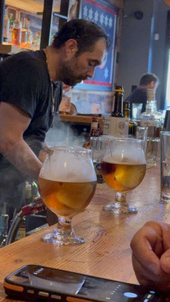 Bartender preparing drinks behind a bar, with two glasses of amber liquid and smoke on the counter. A hand and a smartphone are visible in the foreground.