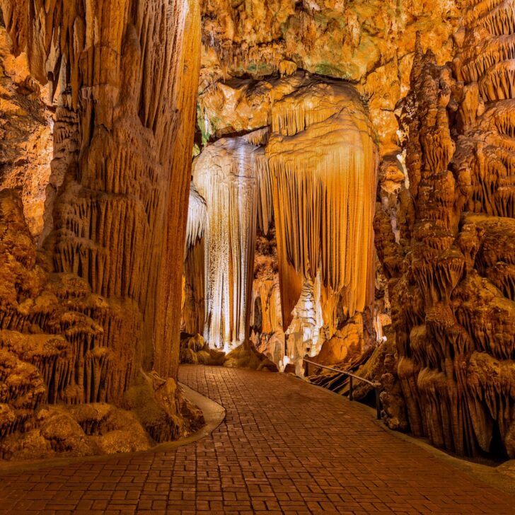 A brick pathway winds through an illuminated cave filled with large stalactites and stalagmites in shades of brown and orange.