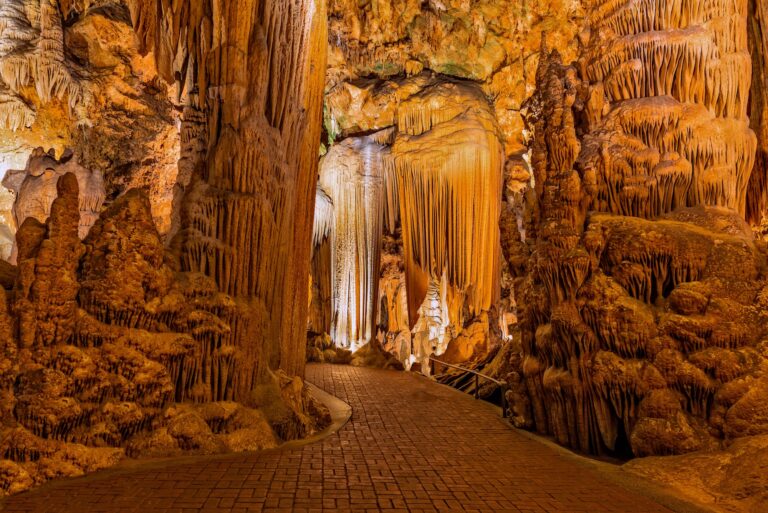 A brick pathway winds through an illuminated cave filled with large stalactites and stalagmites in shades of brown and orange.