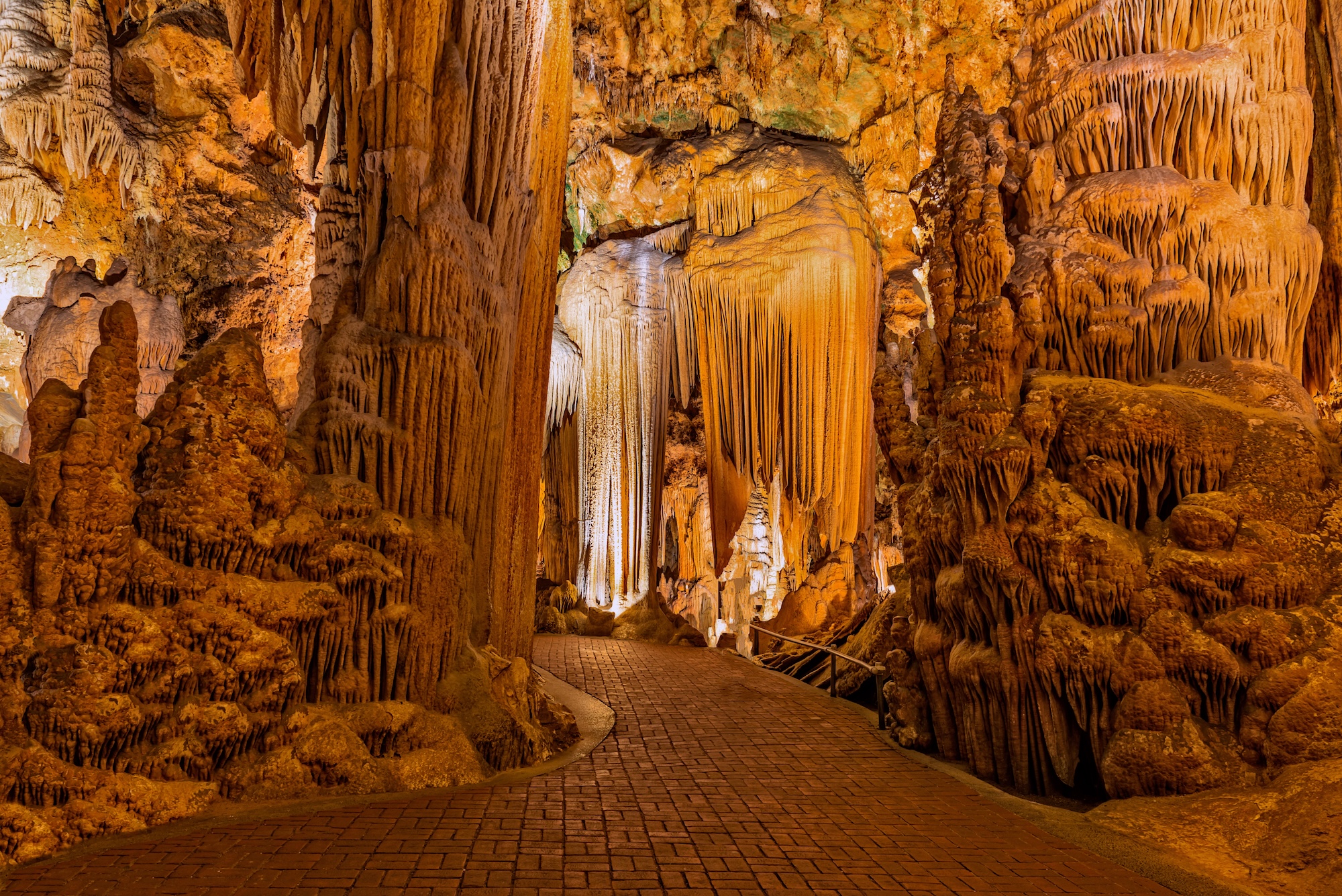 A brick pathway winds through an illuminated cave filled with large stalactites and stalagmites in shades of brown and orange.