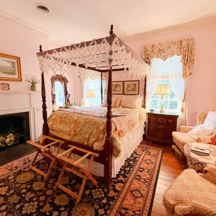 A bright bedroom with a four-poster canopy bed, floral bedding, armchairs, a fireplace, and two windows with curtains. Wooden floors and an ornate rug complete the cozy decor.