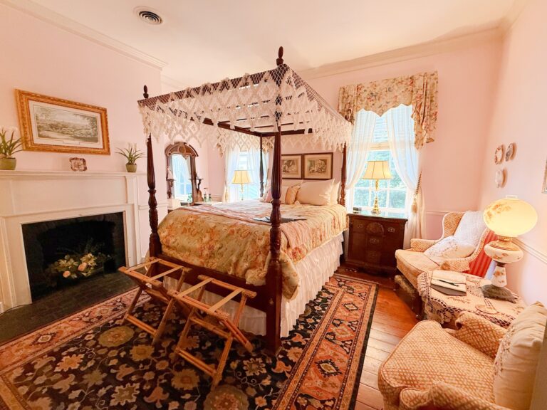 A bright bedroom with a four-poster canopy bed, floral bedding, armchairs, a fireplace, and two windows with curtains. Wooden floors and an ornate rug complete the cozy decor.