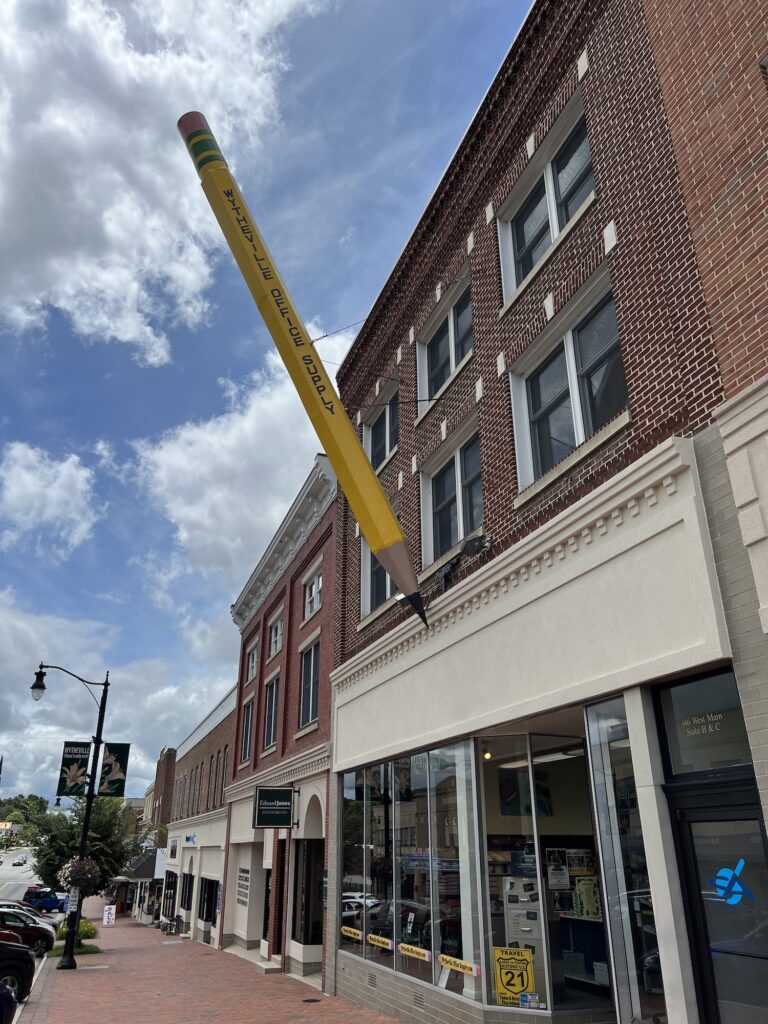 A giant yellow pencil sculpture is attached to the exterior of a brick building on a downtown street under a partly cloudy sky, adding a whimsical touch often found in charming historic Virginia towns.