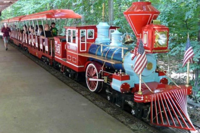 A small red and blue train with American flags is carrying passengers along a track through a wooded area.