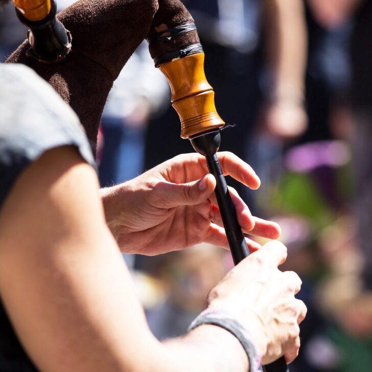 Close-up of a person playing the bagpipes outdoors, with hands on the instrument’s chanter and blurred people in the background.