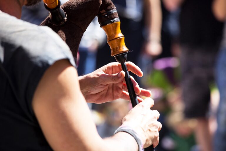 Close-up of a person playing the bagpipes outdoors, with hands on the instrument’s chanter and blurred people in the background.