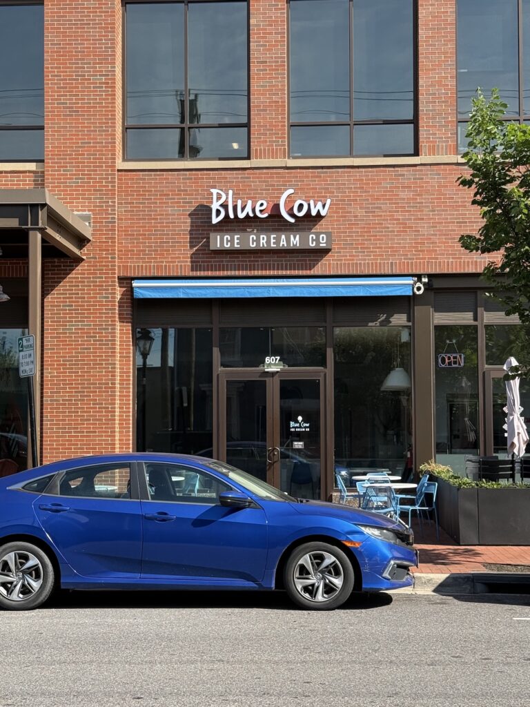 A blue car is parked in front of Blue Cow Ice Cream Co., a brick building with large windows and outdoor seating.