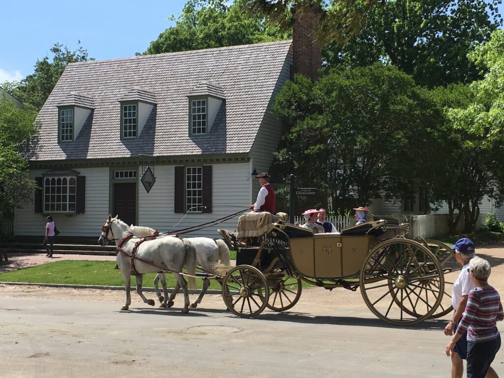 A horse-drawn carriage with people rides past a colonial-style house on a sunny day; others walk nearby.