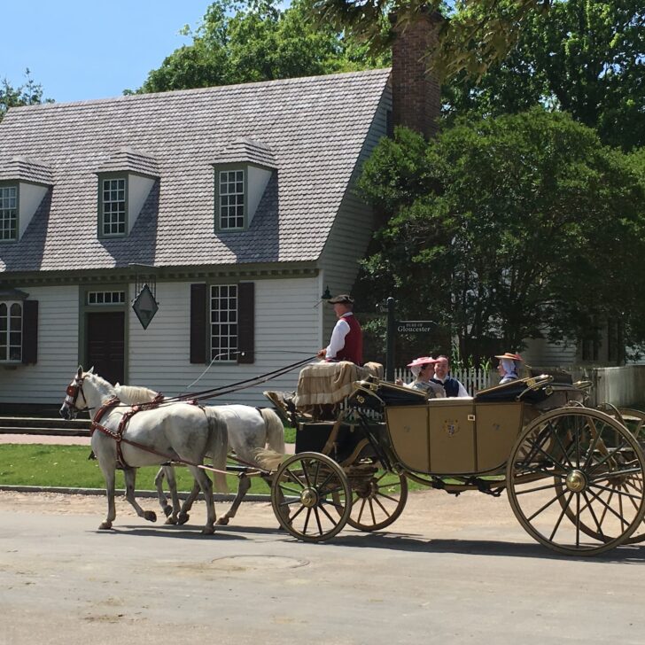 A horse-drawn carriage with people rides past a colonial-style house on a sunny day; others walk nearby.
