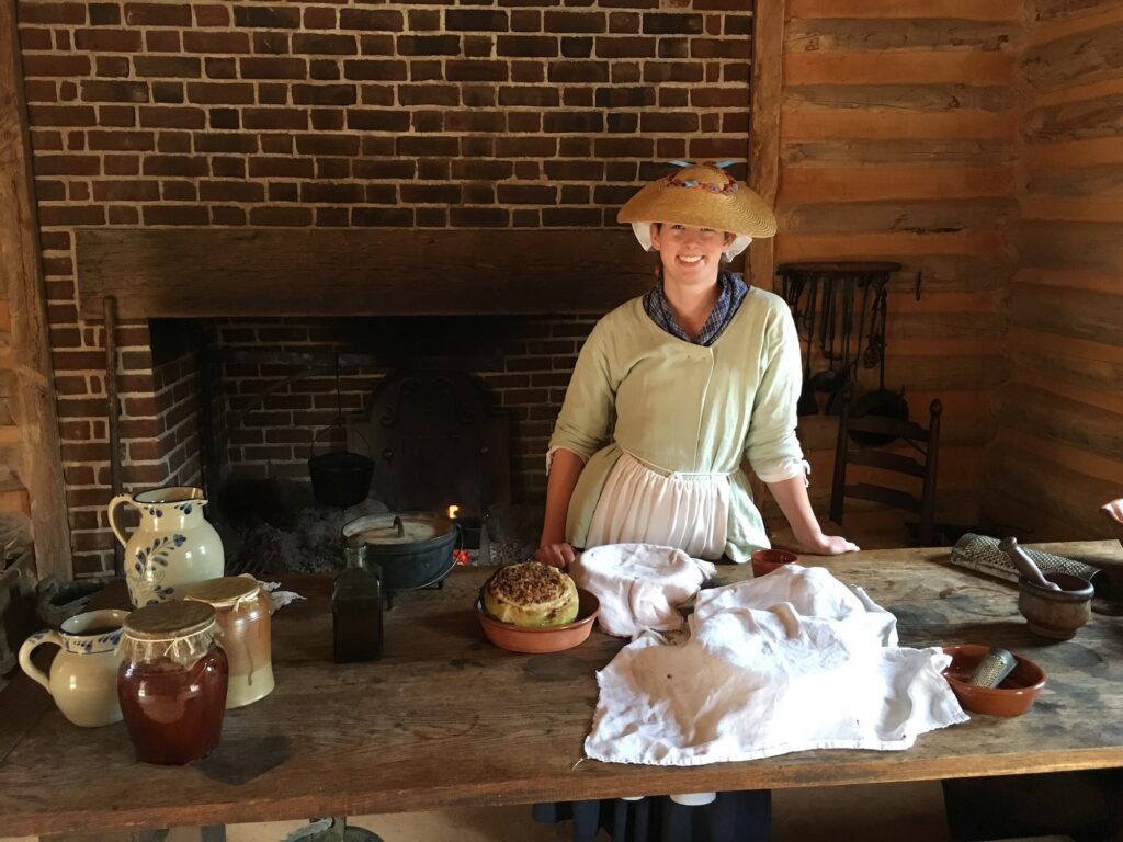 A woman in historical attire stands behind a wooden table with pottery, bread, and kitchen tools in a rustic kitchen with a brick fireplace.