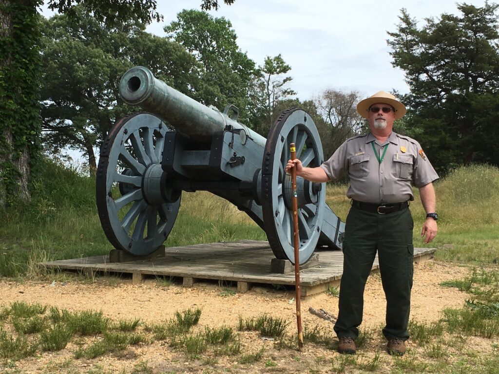 A park ranger stands holding a walking stick next to a large historical cannon mounted on a wooden platform outdoors.