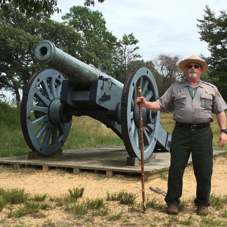 A park ranger stands holding a walking stick next to a large historical cannon mounted on a wooden platform outdoors.
