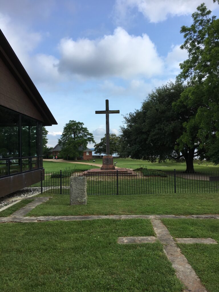 Large stone cross stands within a fenced area on a grassy lawn, with a tree, building, and partly cloudy sky in the background.
