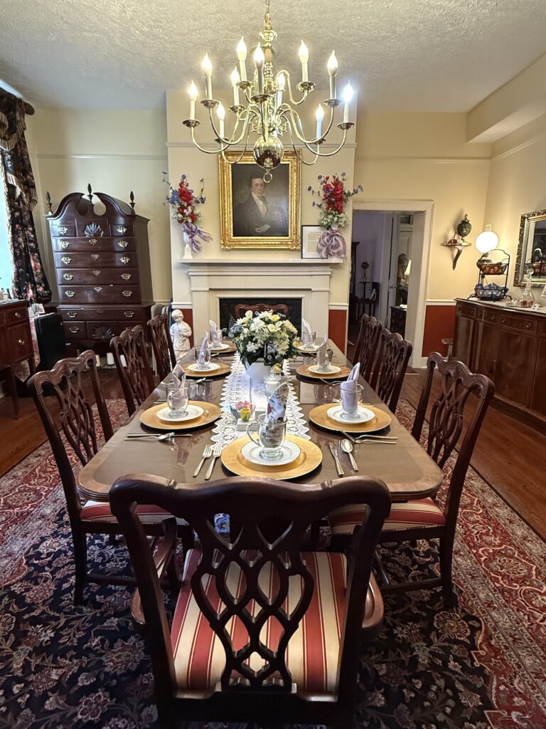 Formal dining room with a set table for eight, ornate wooden chairs, chandelier, floral centerpiece, and a portrait above the fireplace.
