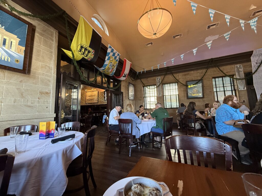 People dine at wooden tables in a German-themed restaurant&mdash;one of the best restaurants in Fredericksburg&mdash;decorated with flags, string lights, and garland. Some tables are empty and others are occupied by groups.