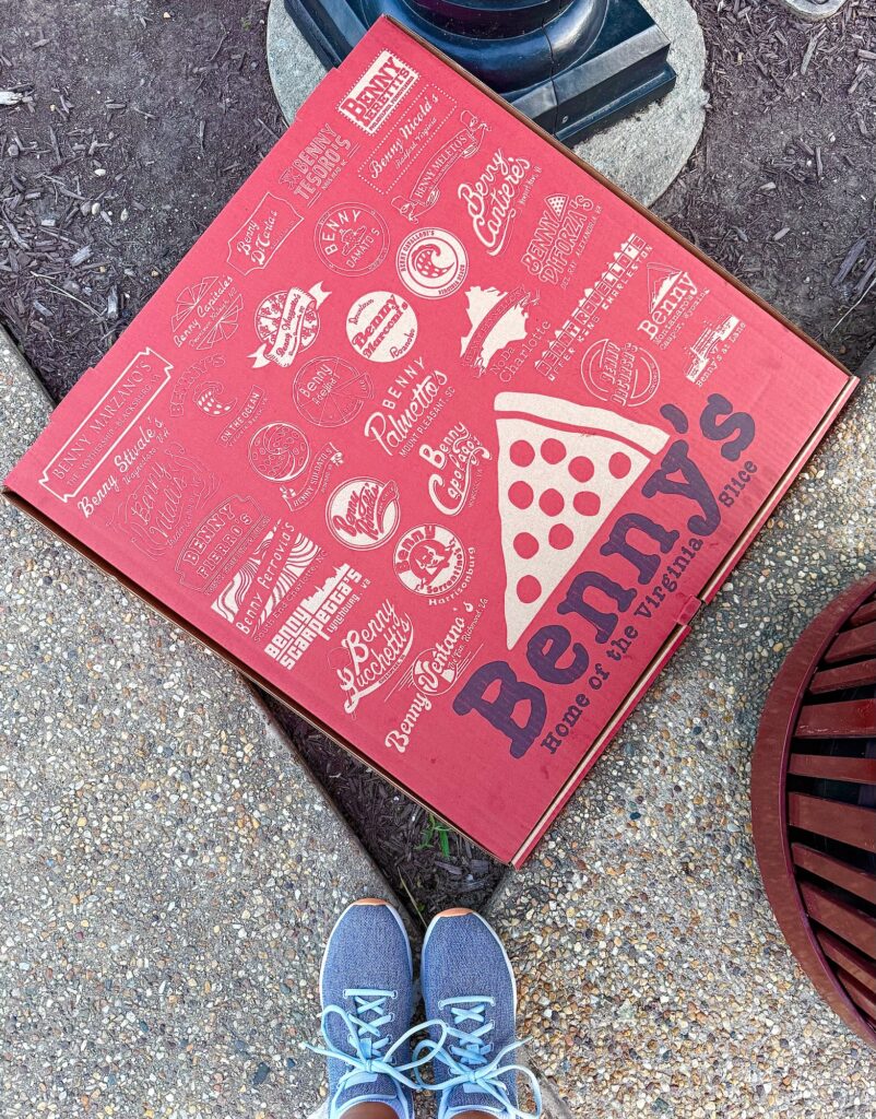 A red Benny&rsquo;s pizza box with logos and a slice graphic sits on the ground near blue sneakers, an outdoor lamp post, and a trash can&mdash;an unexpected scene featuring one of the best restaurants in Fredericksburg.