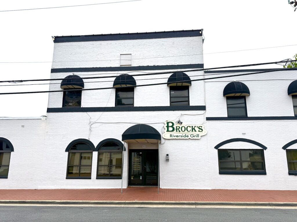 White brick building with black awnings and a sign reading &ldquo;Brock&rsquo;s Riverside Grill&rdquo; above the entrance; view from across the street&mdash;one of the best restaurants in Fredericksburg.