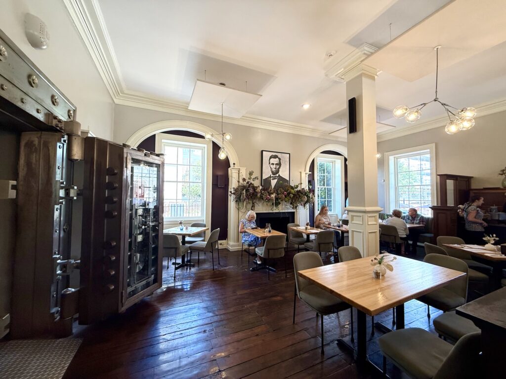 Modern restaurant interior with wooden floors, large windows, a wall mural of Abraham Lincoln, a large open vault, and several people dining at tables—an inviting space that showcases why it’s considered one of the best restaurants in Fredericksburg.
