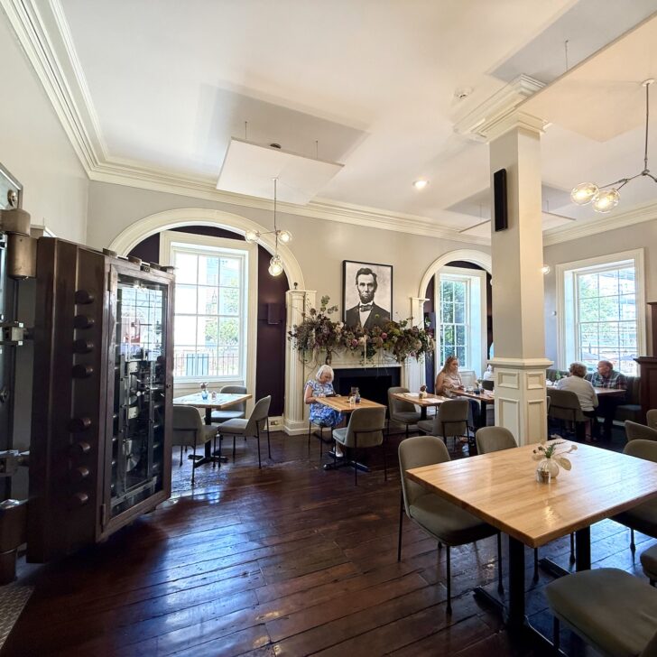 Modern restaurant interior with wooden floors, large windows, a wall mural of Abraham Lincoln, a large open vault, and several people dining at tables—an inviting space that showcases why it’s considered one of the best restaurants in Fredericksburg.