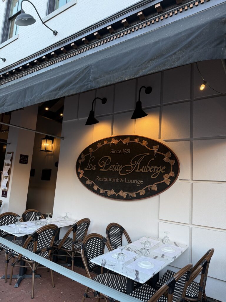 Outdoor seating at La Petite Auberge Restaurant & Lounge, with neatly set tables and chairs beneath a large sign on a white wall, makes it easy to see why this spot is considered one of the best restaurants in Fredericksburg.