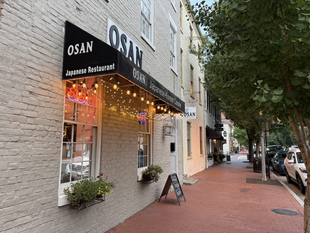Street view of OSAN Japanese Restaurant, celebrated as one of the best restaurants in Fredericksburg, with a black awning, string lights, neon sign in the window, flowers, and a sandwich board on the brick sidewalk.