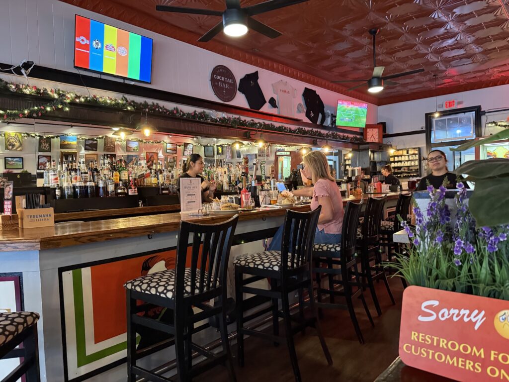 A small group of people sit at a bar inside one of the best restaurants in Fredericksburg, with a red ceiling, wall decor, and a TV displaying colored test bars.
