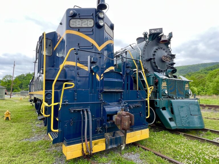 Two vintage trains—a blue and yellow diesel locomotive and a black steam engine—are parked side by side on the tracks, making it a perfect stop for railroad enthusiasts enjoying day trips from Roanoke.