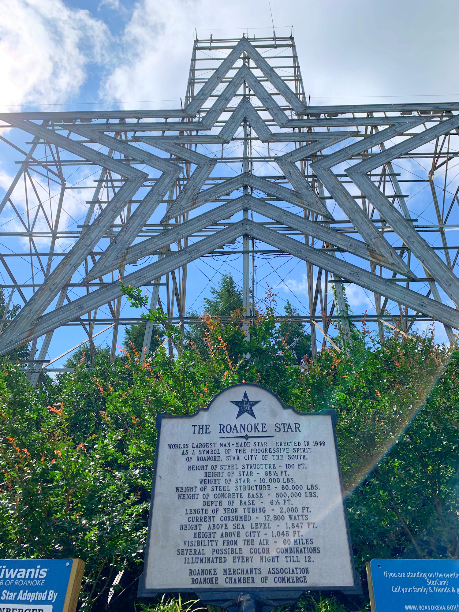 Large metal star structure stands on a hill with a historical marker in front detailing the Roanoke Star’s history, surrounded by greenery and blue sky.